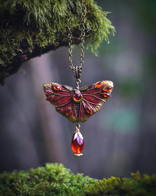 A fantasy-style butterfly pendant necklace with red and gold enamel wings and a dangling faceted amber gem, hanging from a mossy branch in a forest setting.
